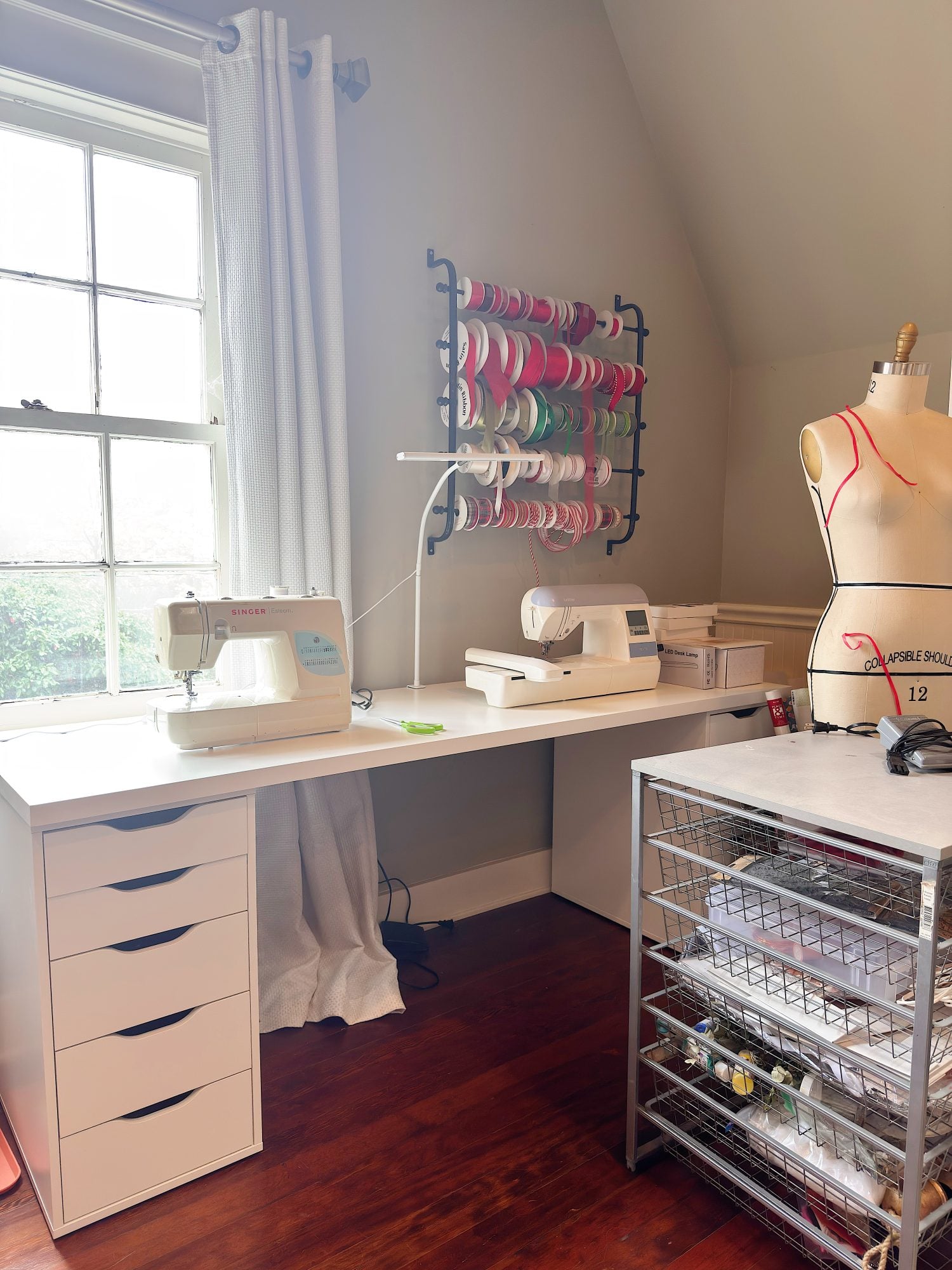 A sewing room with two sewing machines on a white desk, a thread and ribbon rack on the wall, a dress form, and a metal storage cart with organized supplies.