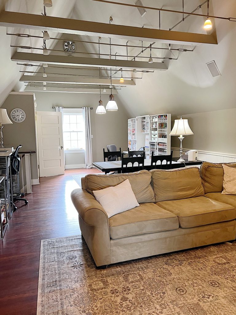 A spacious attic room with a beige sofa, rug, and desk area, featuring vaulted ceilings, exposed beams, hardwood floors, and a window letting in natural light.