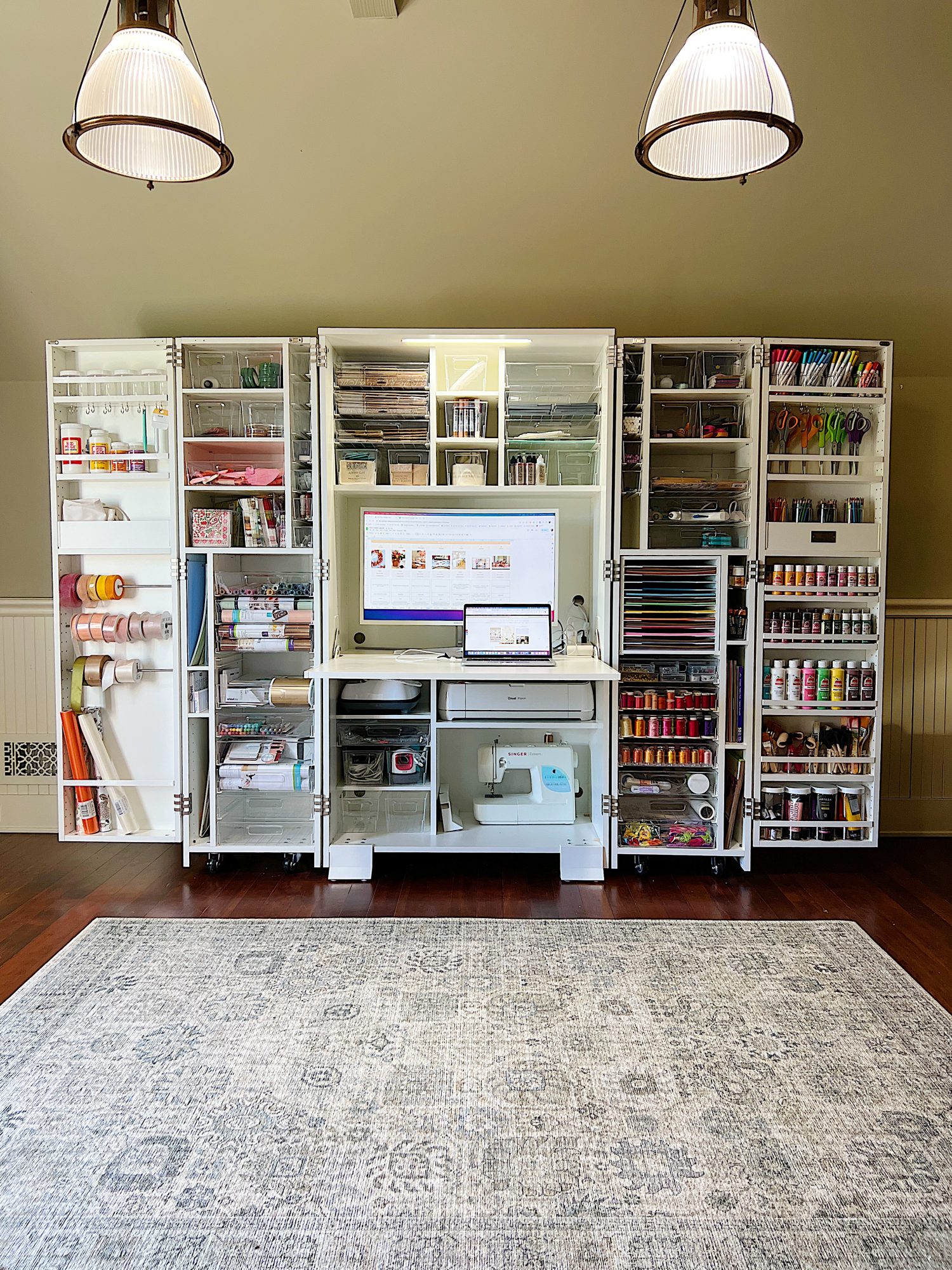 A neatly organized craft station with shelves holding art supplies, a computer monitor, a laptop, a printer, and crafting tools, all set against a white wall with overhead lights.