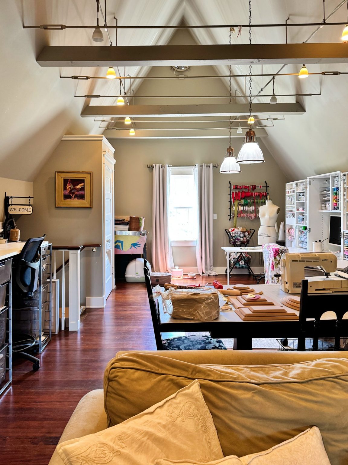 A bright, organized craft room with sewing machines, fabric, storage shelves, a worktable, and a mannequin under high vaulted ceilings with exposed beams.