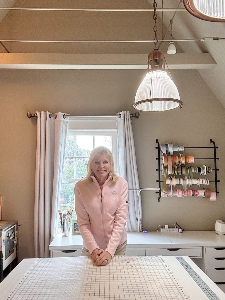 A woman in a pink jacket stands behind a worktable in a craft room with a window, ribbon spools, and pendant lights visible.