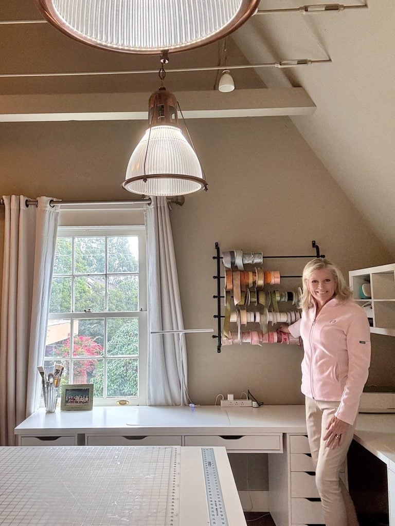 A woman stands in a craft room, holding a measuring stick near a wall-mounted rack of ribbon spools, with a large window and desk in the background.