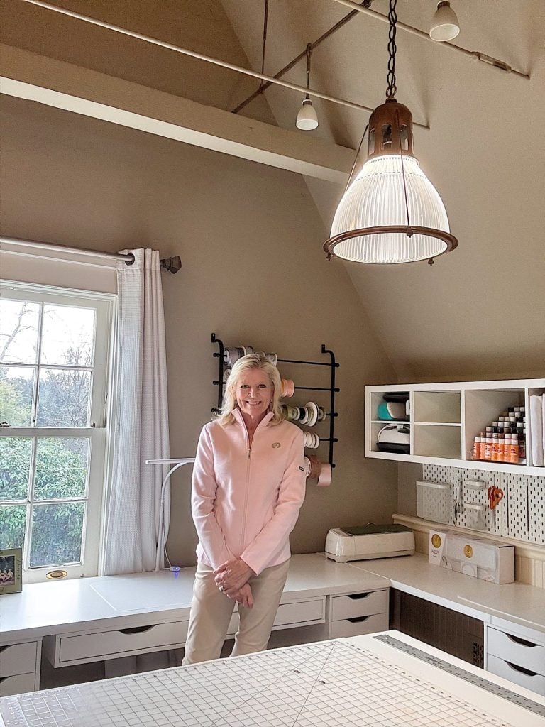 A woman stands and smiles in a craft room with a slanted ceiling, shelves with supplies, a window, and a large overhead light.