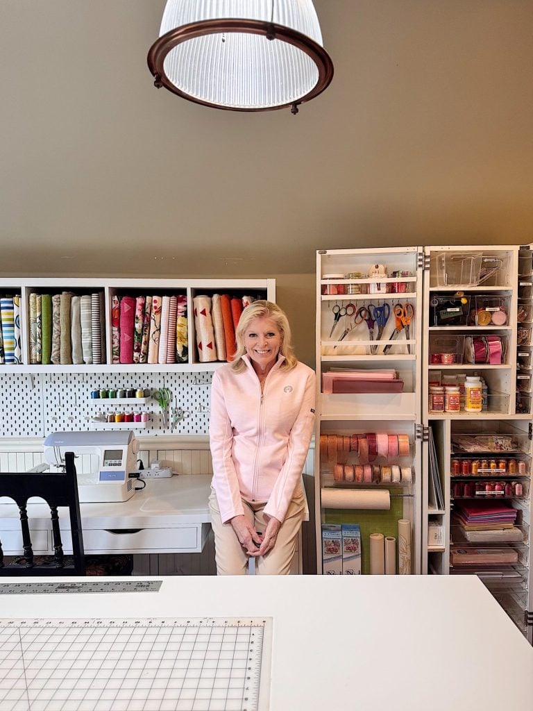 A woman stands in a craft room with shelves of fabric, ribbons, and supplies, next to a sewing machine and a worktable with a cutting mat under a hanging light.