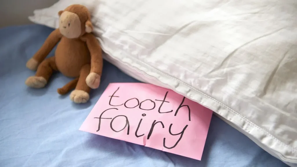 A pink note labeled "tooth fairy" is partially tucked under a white pillow on a blue bed, with a brown stuffed monkey beside it.