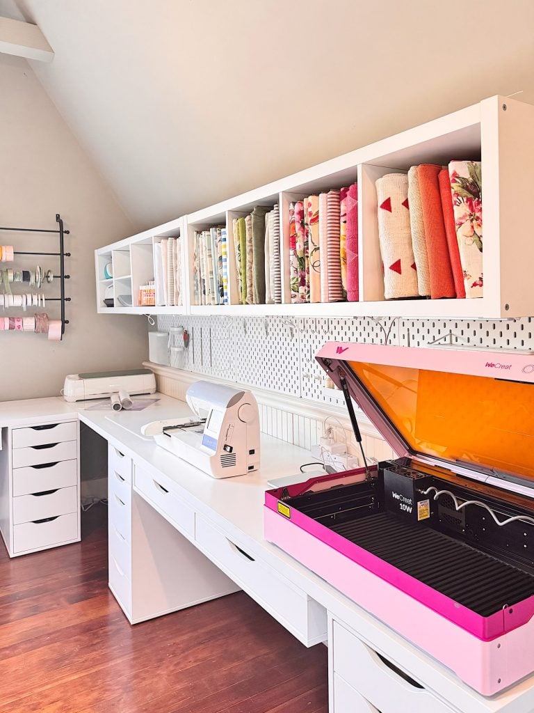 A bright craft room with organized shelves holding fabric, a sewing machine on a white desk, and a pink-lidded cutting machine on the countertop.