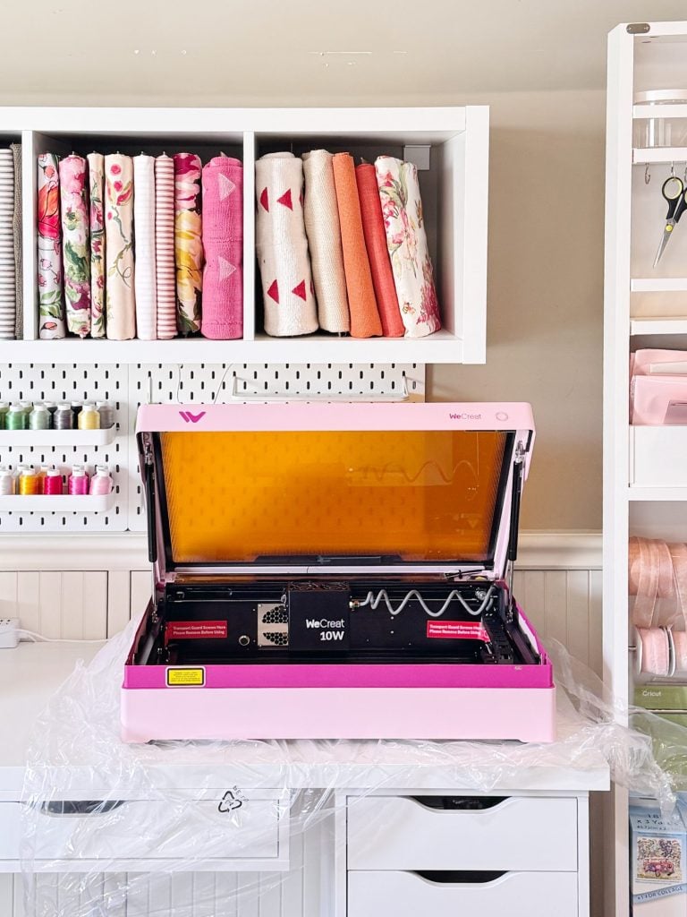 A pink laser cutting machine sits open on a desk, with fabric, thread, and craft supplies organized on shelves in the background.