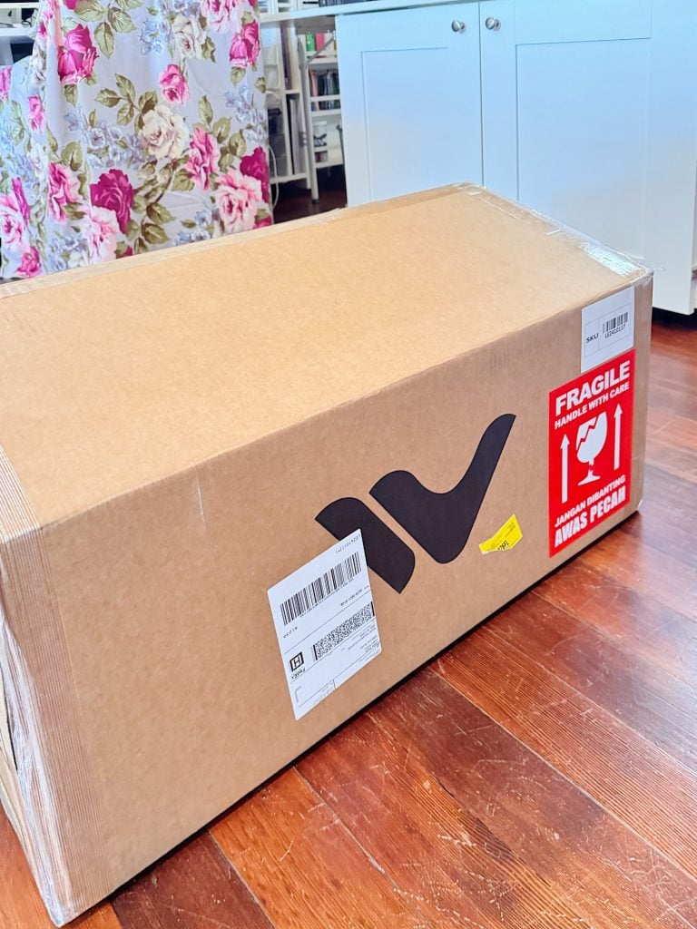 A large cardboard box with a shipping label and a red "fragile" sticker sits on a wooden floor in a bright room with floral and white furniture.