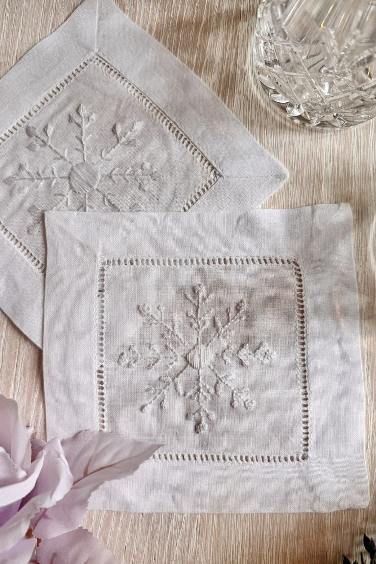 Two white linen napkins with embroidered snowflake designs are placed on a light wooden surface, next to a glass and a pale pink flower.
