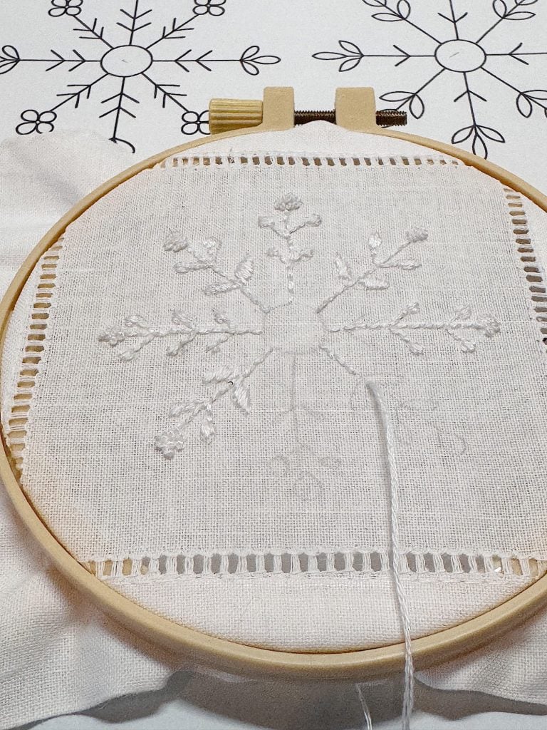 An embroidery hoop holds white fabric with an unfinished snowflake design being stitched; a similar snowflake pattern is printed on paper below.