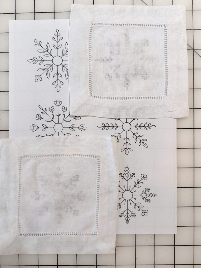 Two pieces of white fabric with embroidered snowflake patterns are placed over printed snowflake designs on a gridded surface.