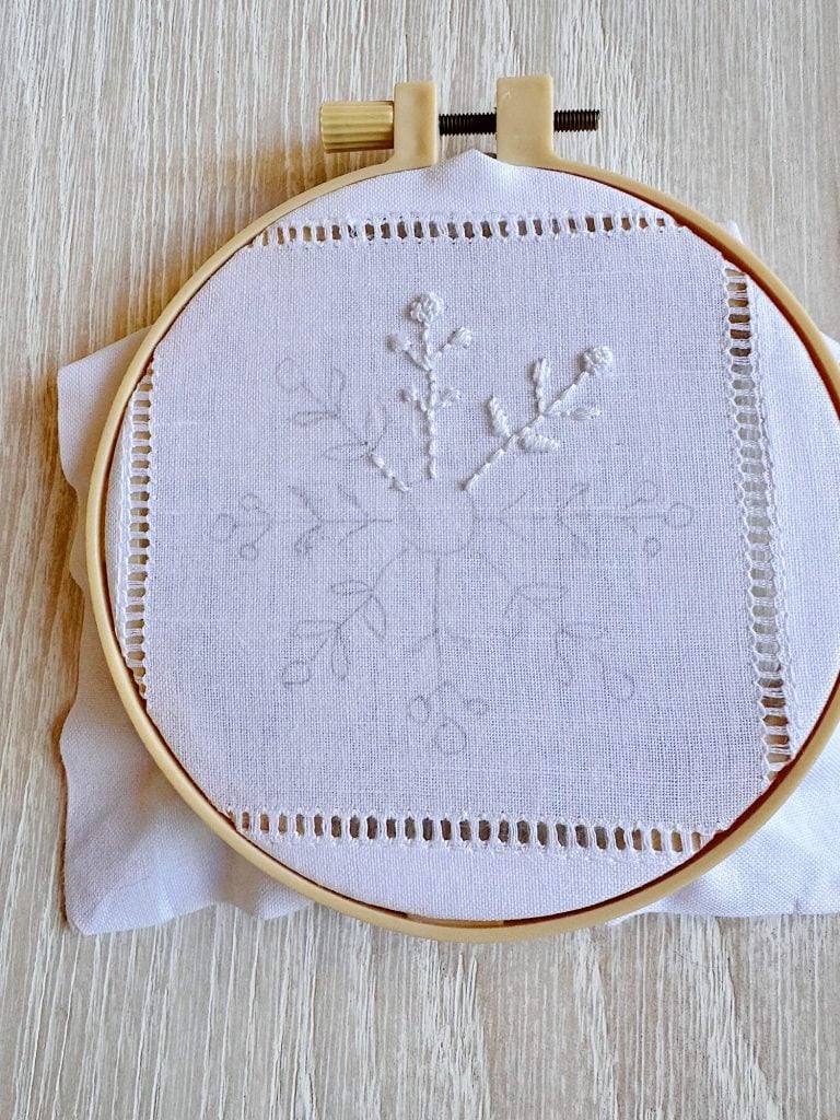 An embroidery hoop holds a piece of white fabric featuring a delicate, partially finished floral or snowflake pattern stitched in white thread.