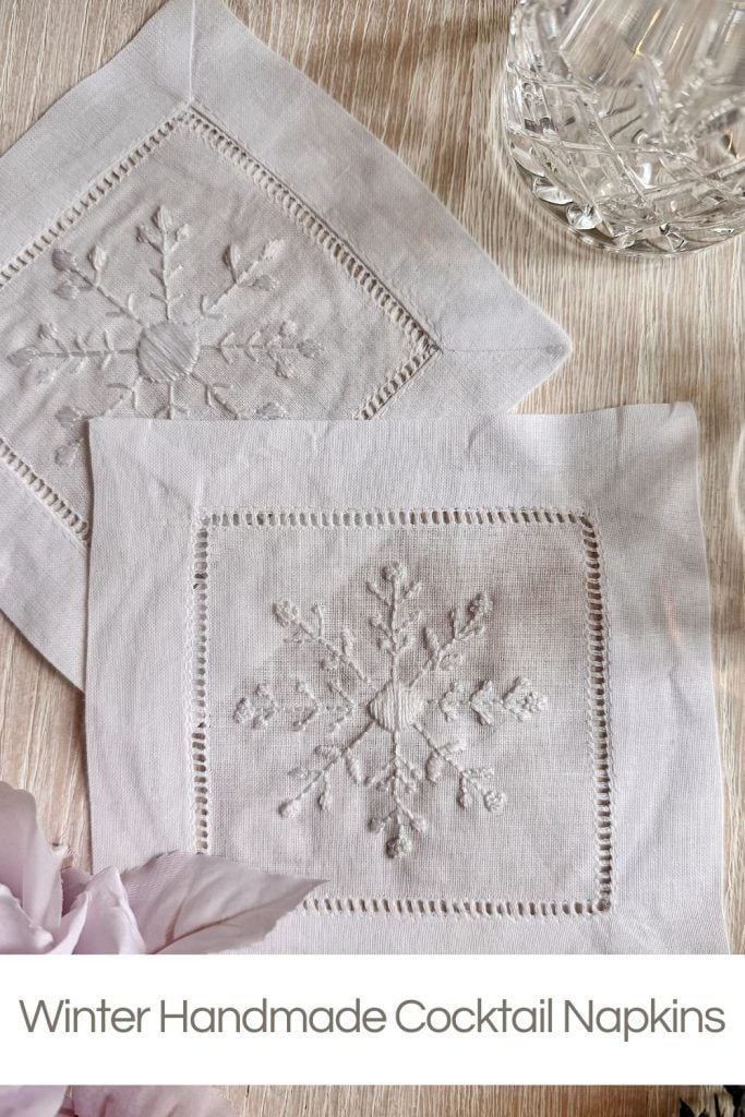 Two white linen cocktail napkins with embroidered snowflake designs are displayed on a light surface next to a crystal glass and pale flowers.
