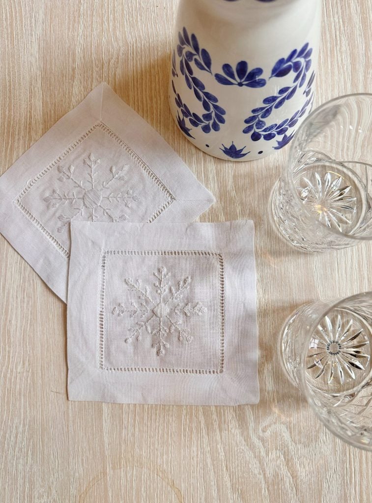 Two embroidered white linen coasters on a light wooden table, next to a white and blue ceramic pitcher and two crystal glasses.