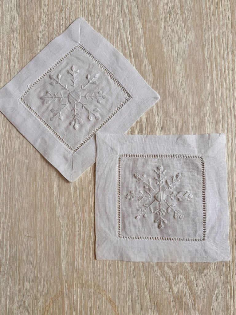 Two white cloth napkins with embroidered snowflake designs, placed on a light wooden surface.