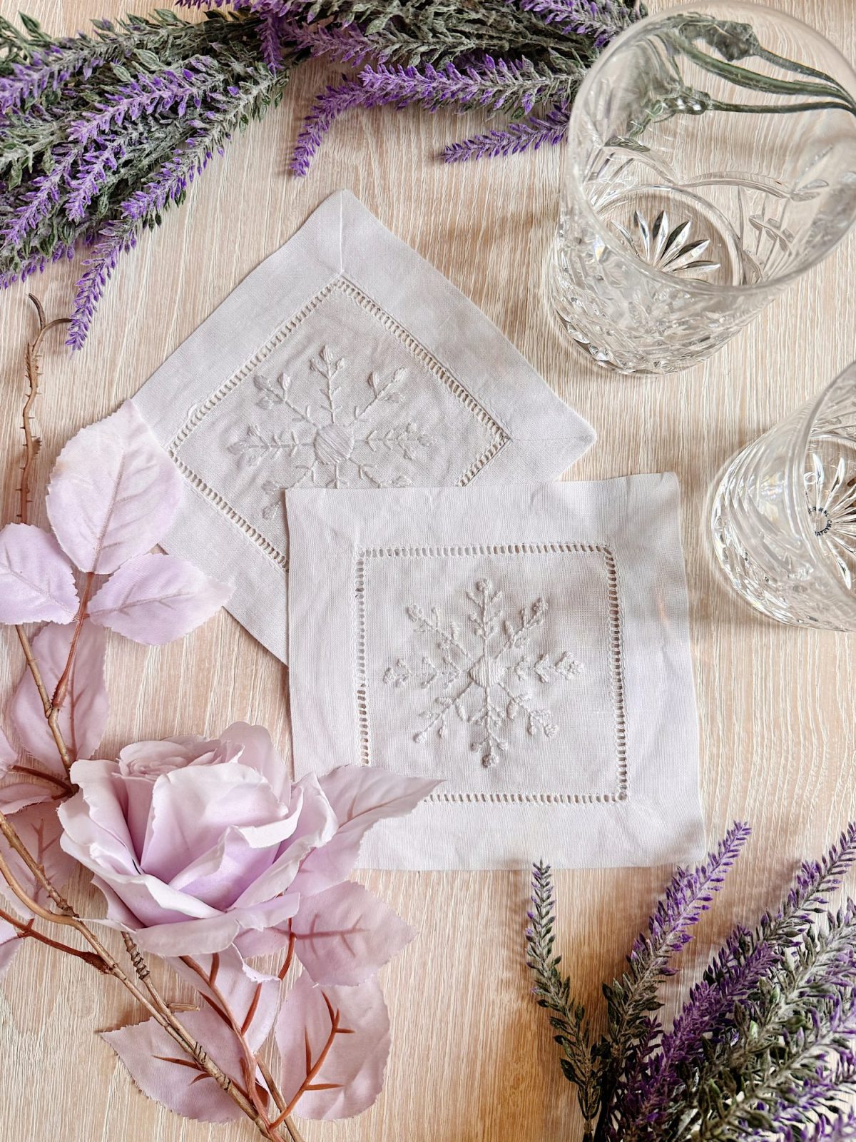 Two clear crystal glasses and two embroidered white napkins with snowflake designs are arranged on a light wooden surface with lavender sprigs and a light purple artificial rose.