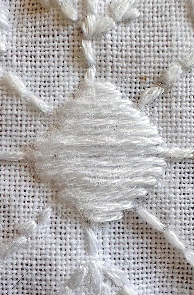 Close-up of white thread embroidered in a geometric pattern on white fabric with visible textured weave and stitching details.