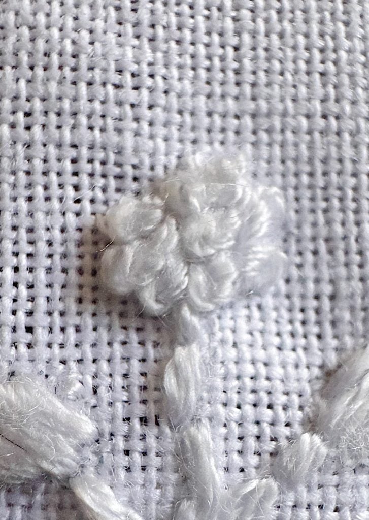 Close-up of white embroidery on white fabric, featuring a cluster of knotted stitches forming a small flower shape with a stem.