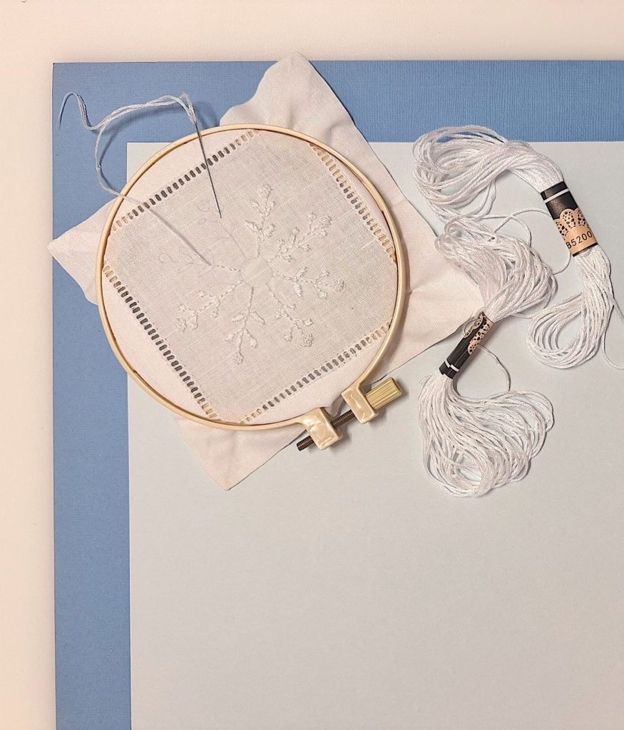 Embroidery hoop with white fabric featuring a stitched snowflake, placed next to white embroidery floss and a needle on a blue and white background.