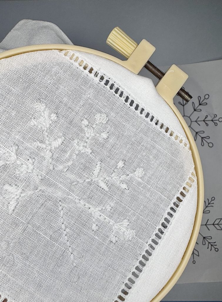 A close-up of white floral embroidery in progress on a piece of fabric stretched in an embroidery hoop, with a floral pattern guide visible beside it.