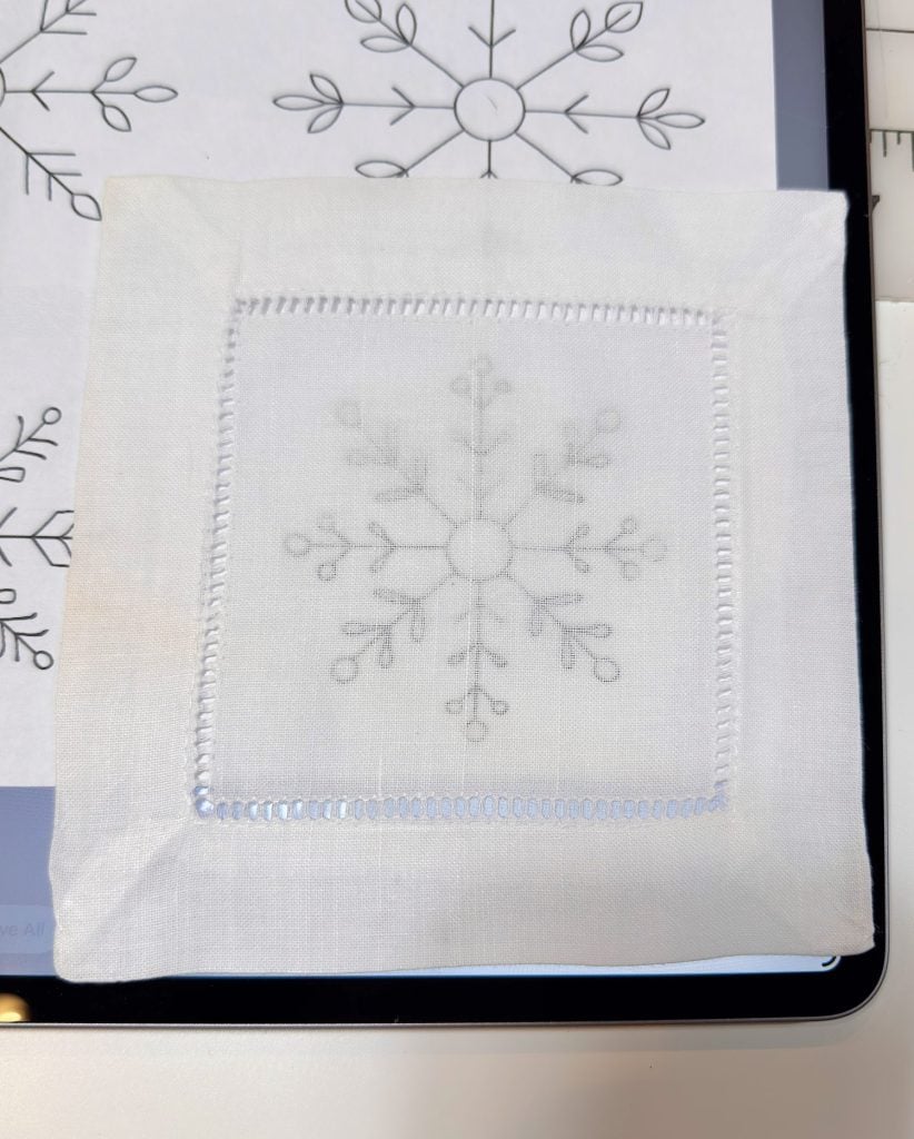 A square piece of white fabric with a hand-drawn snowflake pattern is placed on top of a printed snowflake design.