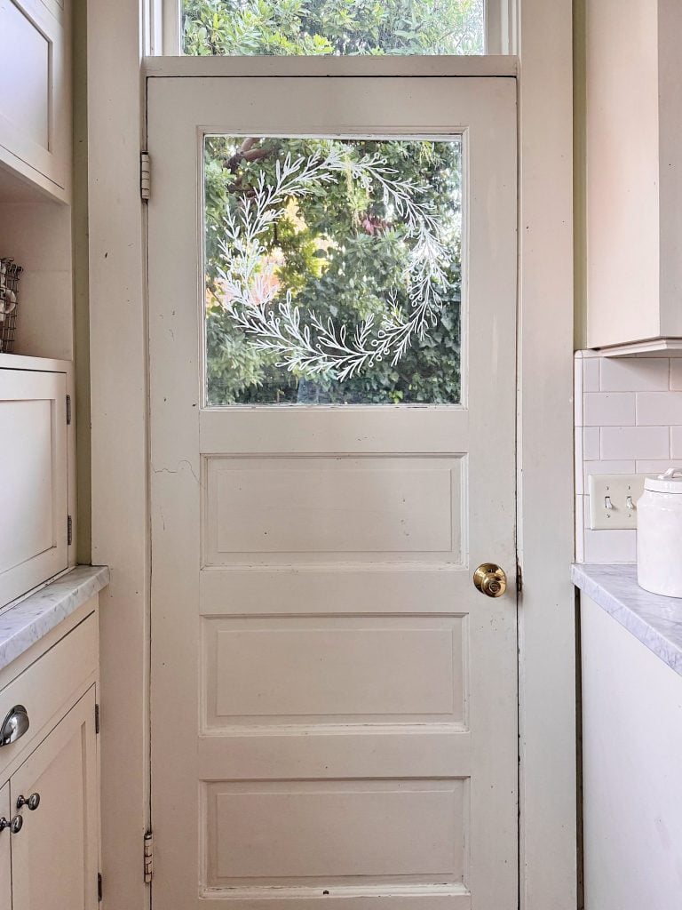 A white wooden door with a glass window featuring a painted leaf wreath design, set in a bright kitchen with white cabinets and countertops.