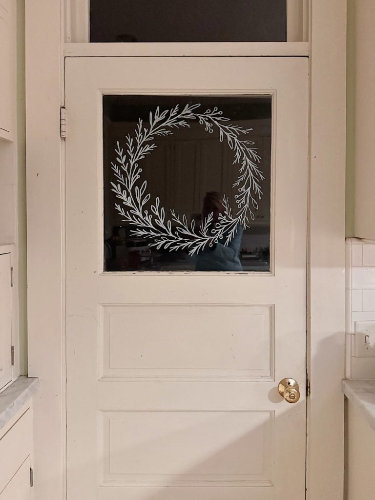 A white door with a glass panel decorated with a hand-drawn white wreath; a person&rsquo;s reflection is faintly visible in the glass.
