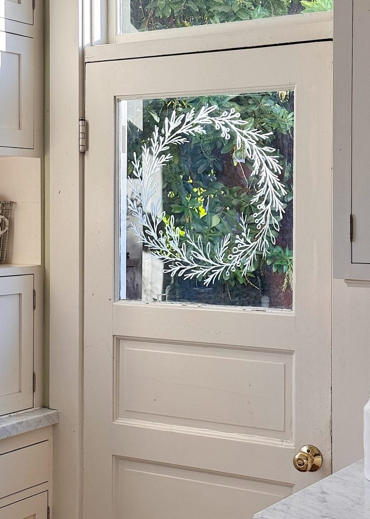 A white wooden door with a glass panel decorated with a white painted wreath design, set in a kitchen with white cabinets and greenery visible outside.