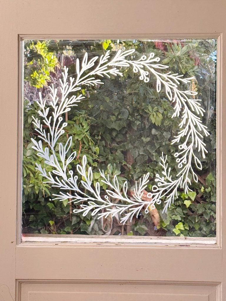 A clear glass window with a white painted wreath design, showing green foliage and plants outside.