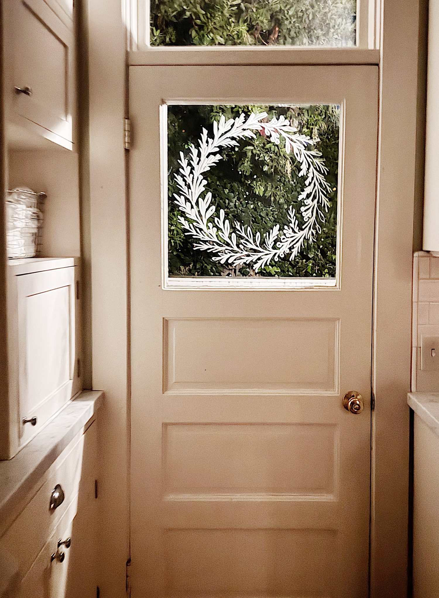 A beige door with a glass window features a white painted wreath design, seen from inside a kitchen with cabinets and a countertop nearby. Trees are visible through the window.
