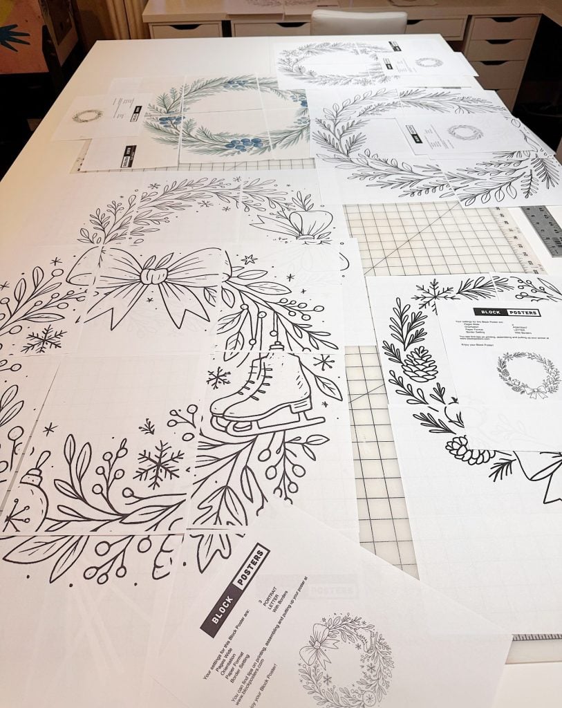 Large sheets of paper with black and white holiday wreath designs, including bows and ice skates, are spread out on a work table with rulers and additional printed sheets.