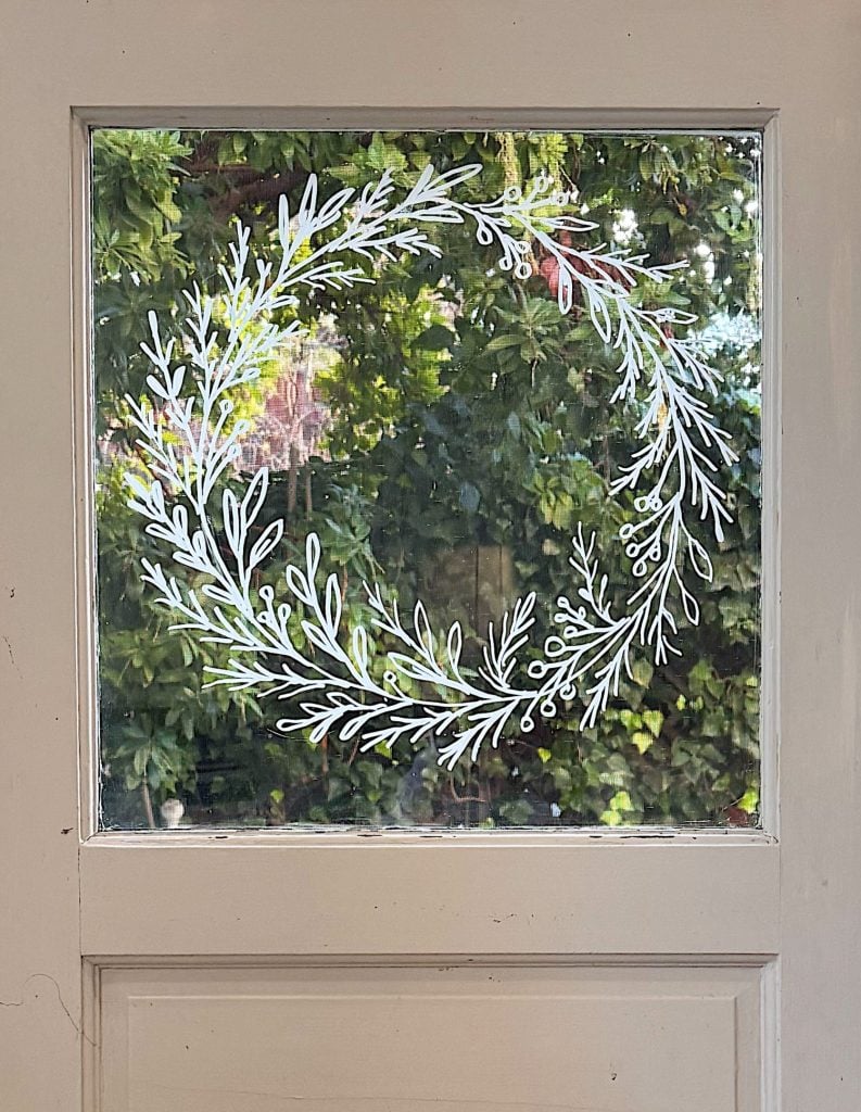 A white, hand-drawn wreath design decorates a windowpane on a beige door, with green foliage visible outside.