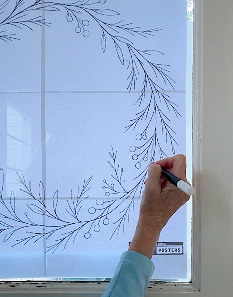 A person traces a botanical wreath design on paper taped to a window, holding a black marker in their right hand.