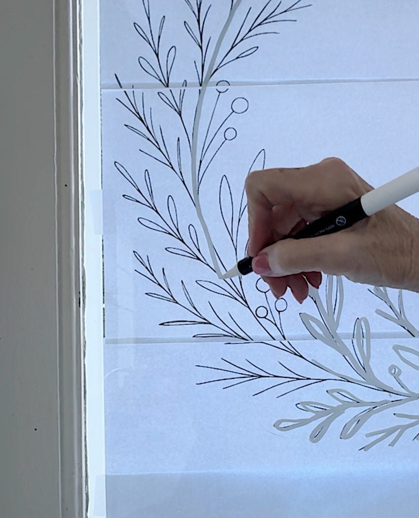 A hand is tracing a leaf and berry design onto a window using a black marker.