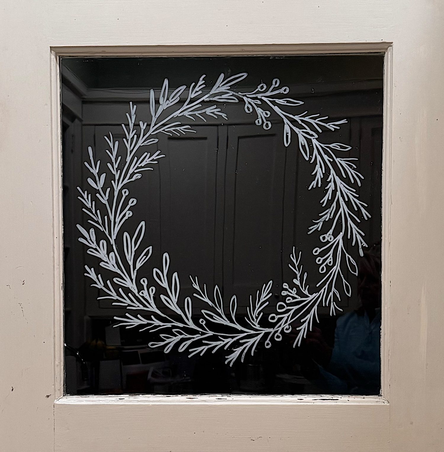 A simple white wreath design is drawn on a glass windowpane set in a beige door, with a faint reflection visible in the glass.