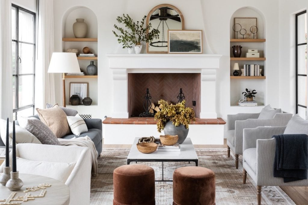 A modern living room with a white fireplace, a round mirror, neutral-toned furniture, built-in shelves, and decorative vases and plants.