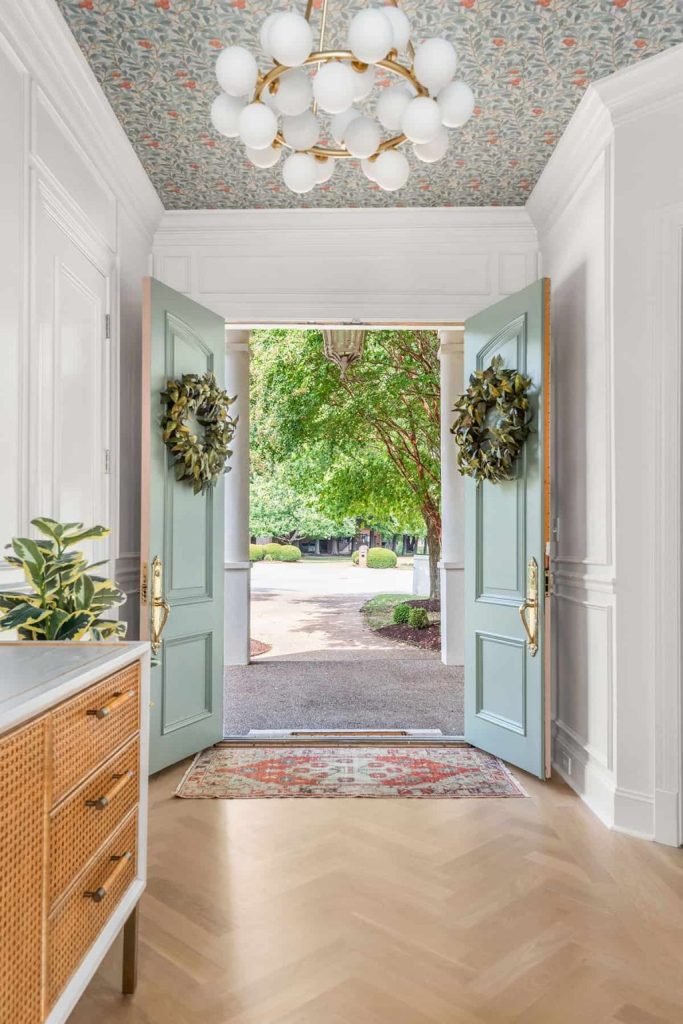 A double-door entryway with green wreaths opens to a driveway and trees. The space features a floral ceiling, light fixture, dresser, and patterned rug on herringbone wood floor.