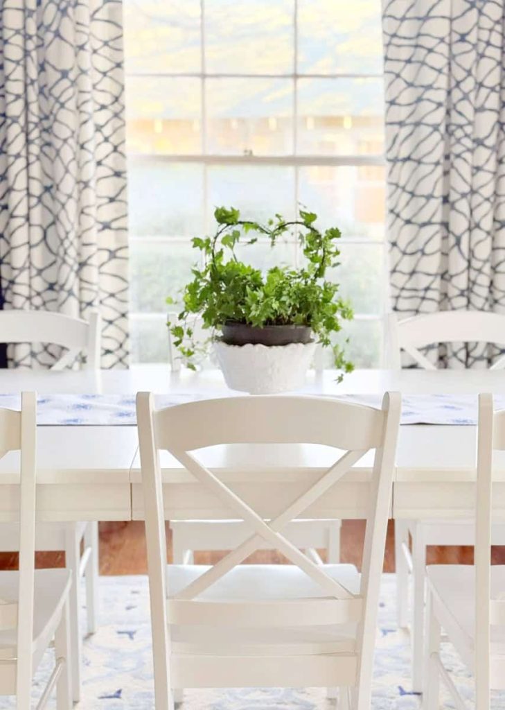 A white dining table with white chairs and a potted green plant in the center, set in front of a large window with patterned curtains.