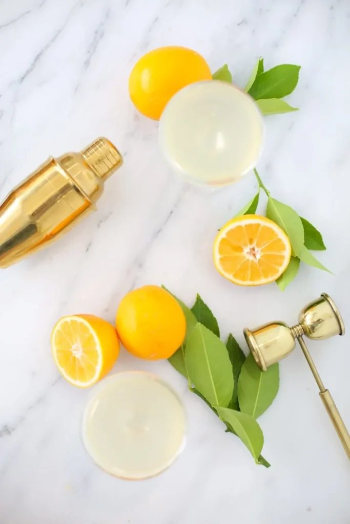 A gold cocktail shaker, a jigger, fresh lemons, lemon halves, green leaves, and two filled cocktail glasses arranged on a white marble surface.
