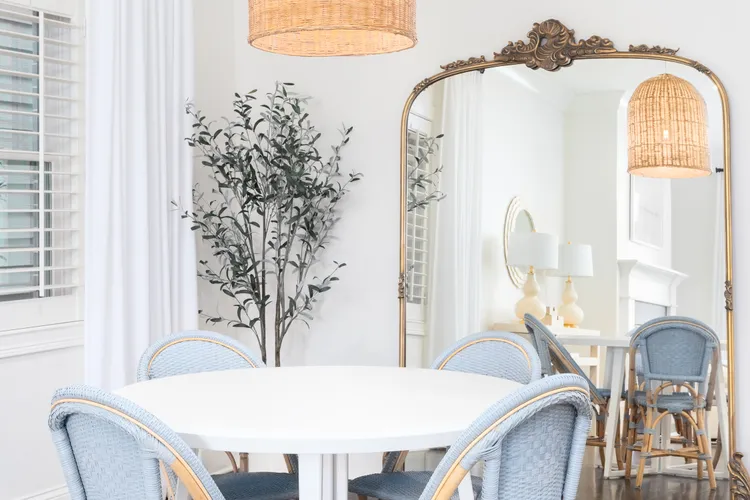 A round white dining table with light blue chairs sits in front of a large ornate mirror, with a potted plant and a pendant light visible in a bright, airy room.