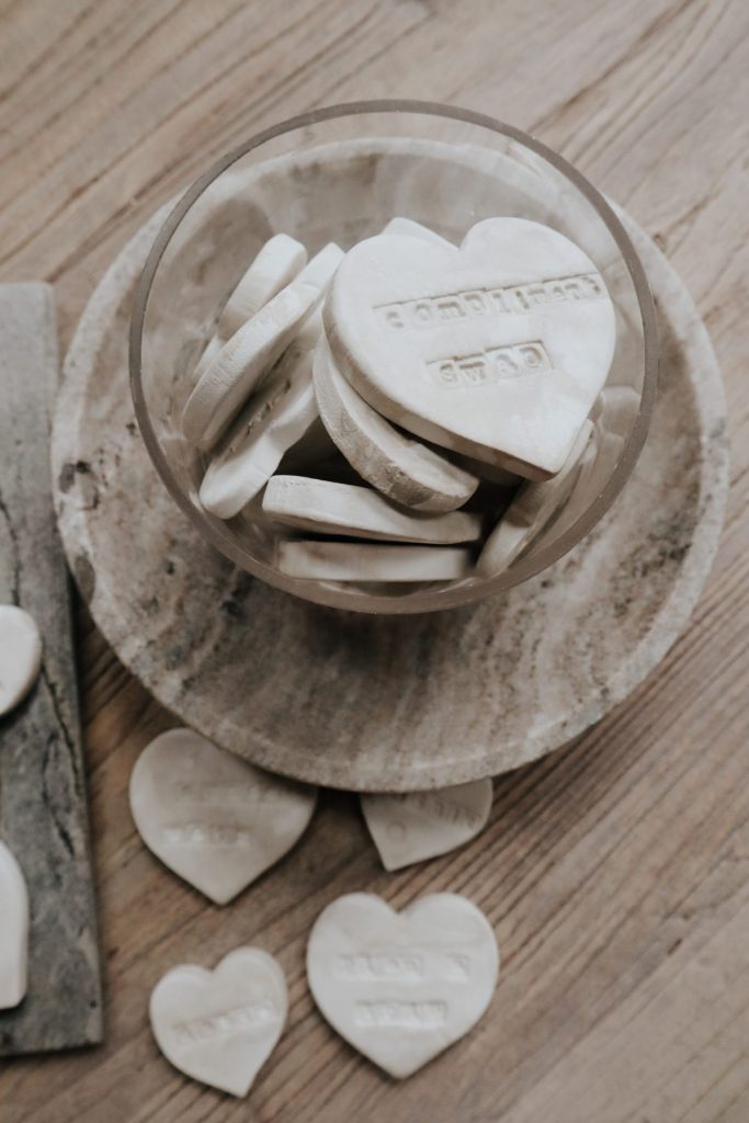 A glass bowl filled with white, heart-shaped cookies or candies sits on a round marble plate, with more heart-shaped pieces scattered nearby.