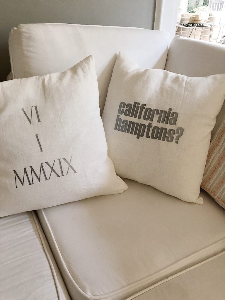 Two white throw pillows on a white sofa; one has "VI I MMXIX" and the other reads "california hamptons?" in gray text.