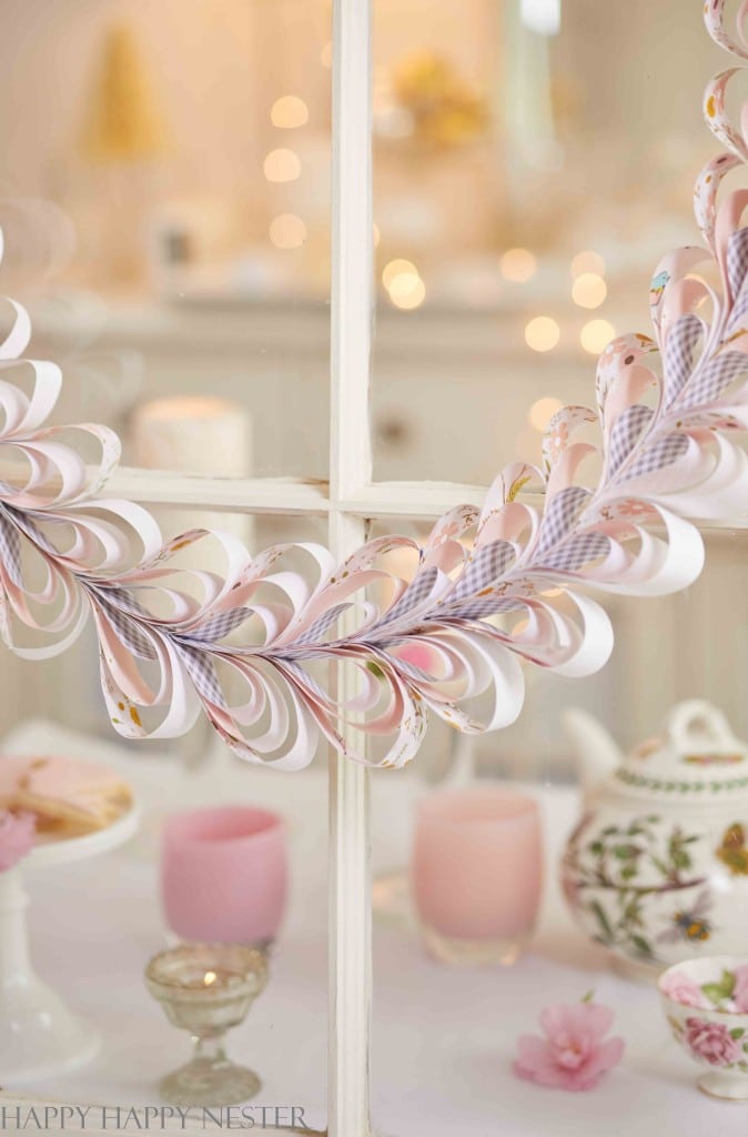 A paper heart garland hangs on a windowpane with a teapot, pink candles, and blurred decorative items in the background.