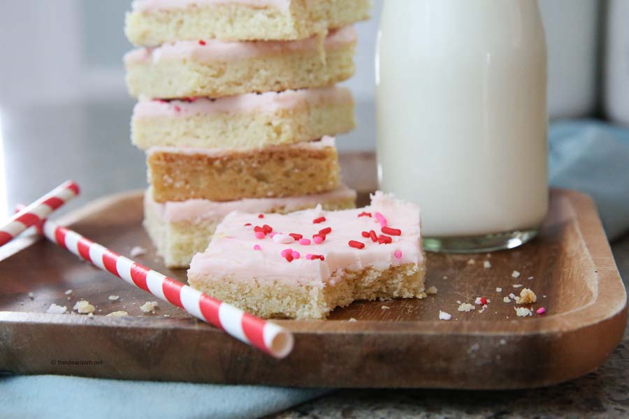 A wooden tray with frosted sugar cookie bars, red and pink heart sprinkles, two striped straws, and a bottle of milk.