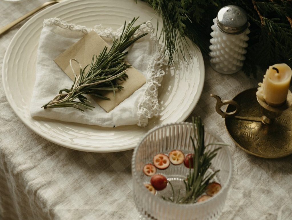 A table setting with a white plate, cloth napkin, and rosemary sprig, next to a glass with cranberries, rosemary, a brass candle holder, and a salt shaker on a linen tablecloth.