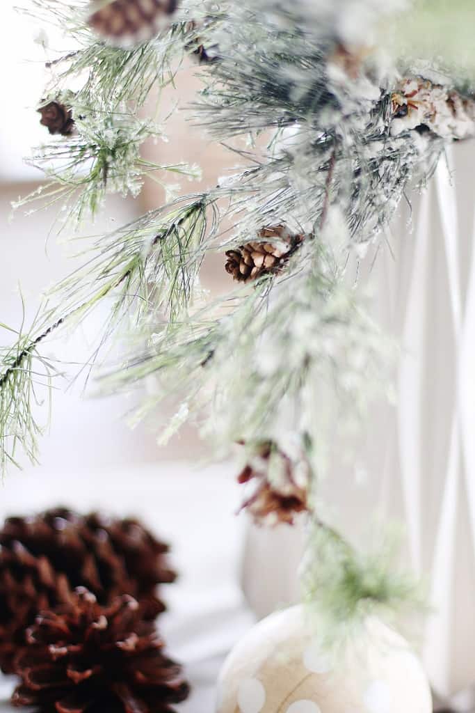 Frosted pine branches with small pinecones hang above a surface with a large pinecone and a beige ornament with white polka dots.