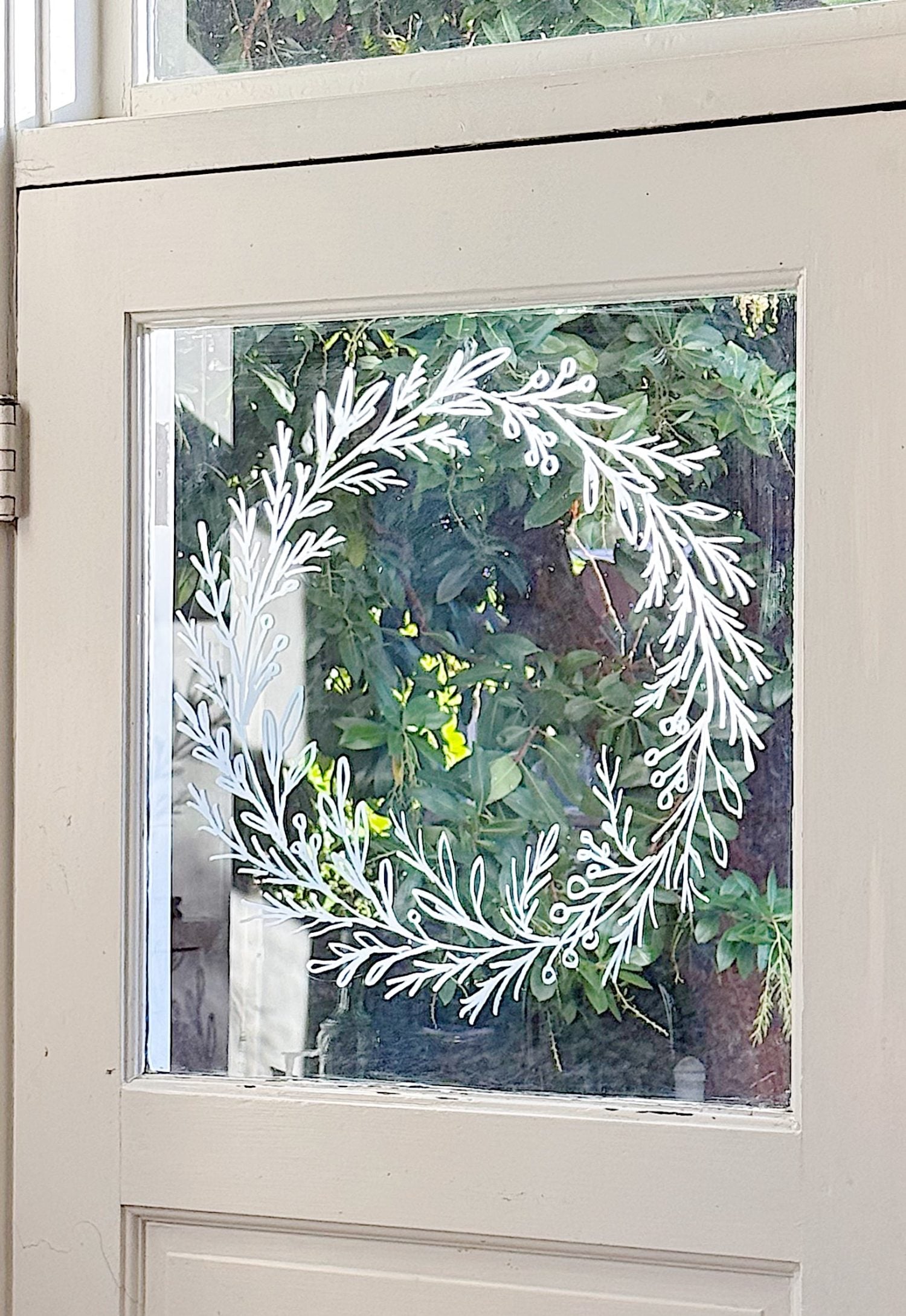 A white leafy wreath design is drawn on a glass windowpane of a beige door, with green foliage visible outside.