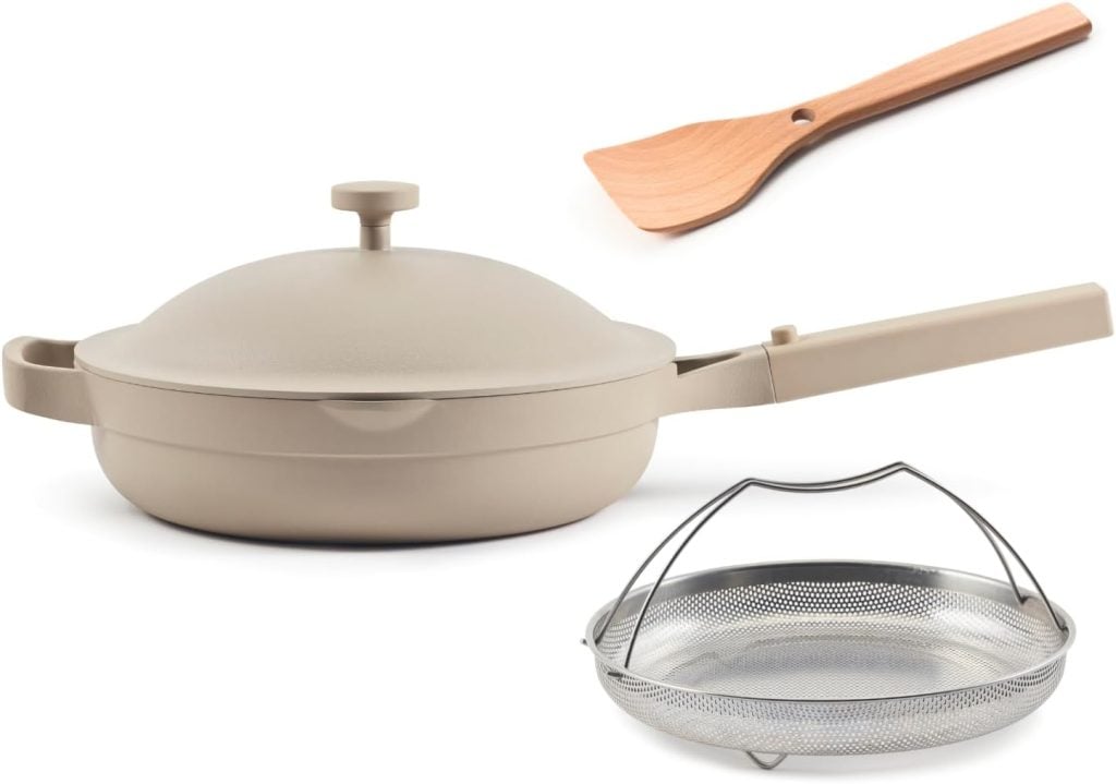 A beige nonstick pan with lid, a wooden spatula, and a metal steamer basket, all displayed on a white background.