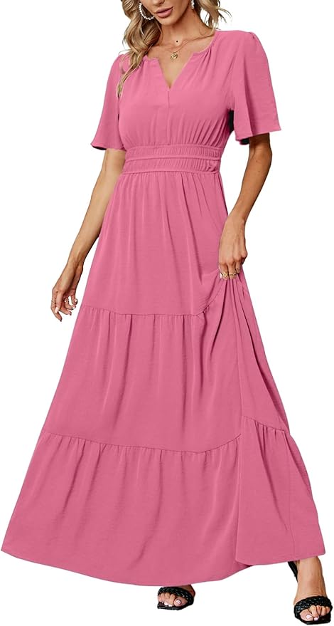 A woman wears a short-sleeve, V-neck, ankle-length pink maxi dress with a fitted waist and tiered skirt, paired with black sandals and gold jewelry.
