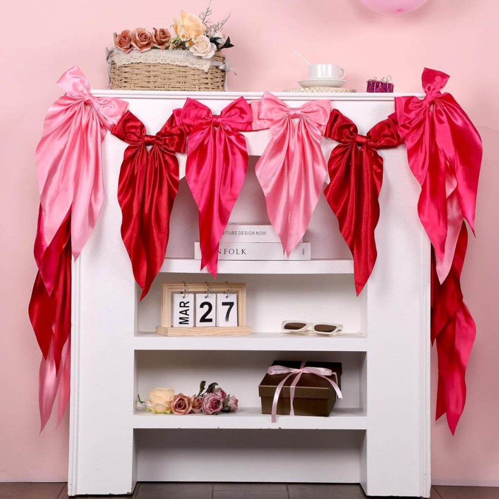 A white mantel decorated with large satin bows in shades of pink and red, with flowers, books, sunglasses, a gift box, and a calendar displaying March 27 below.